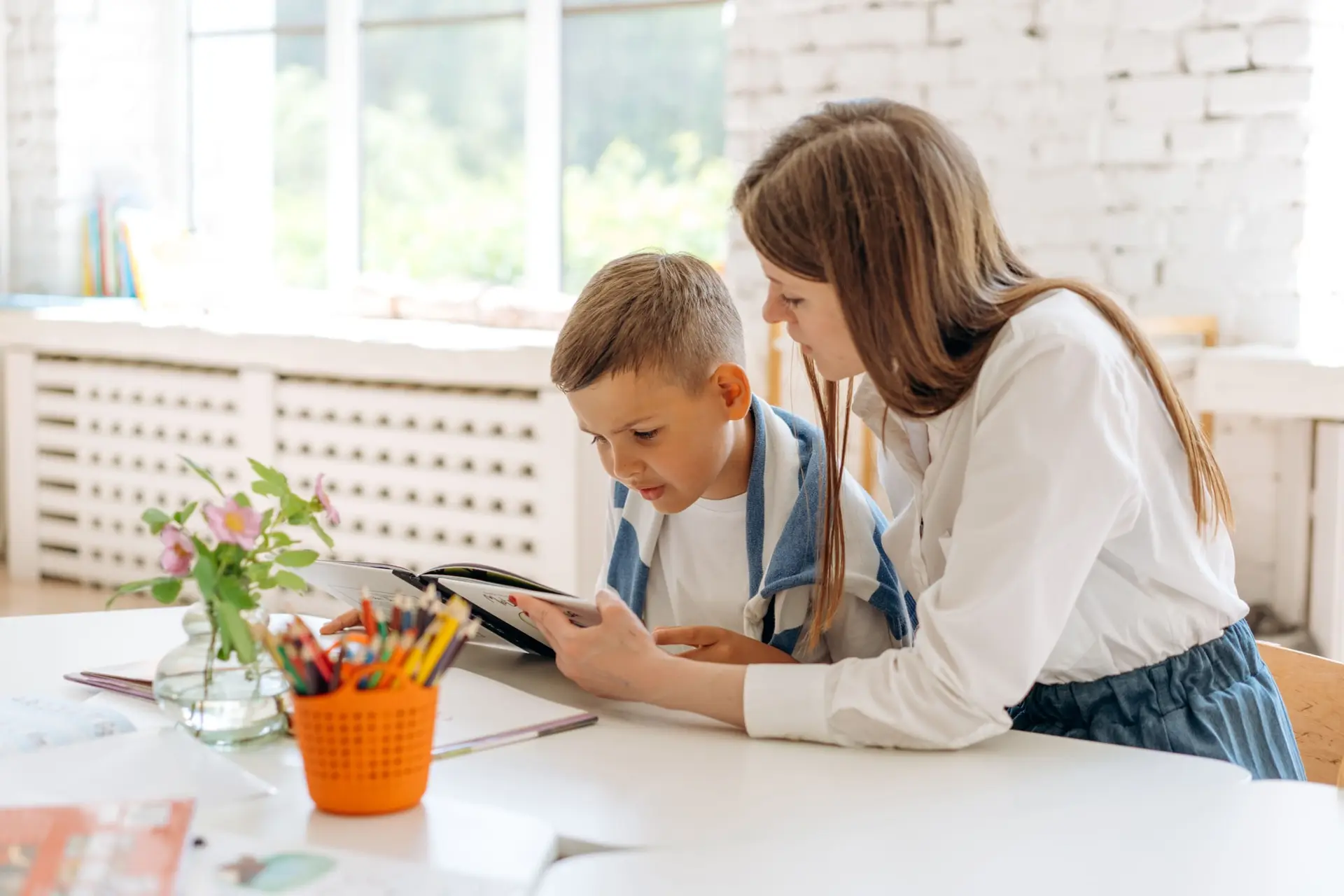 Una adulta y un niño revisan juntos una tableta sobre una mesa con flores y lápices de colores.