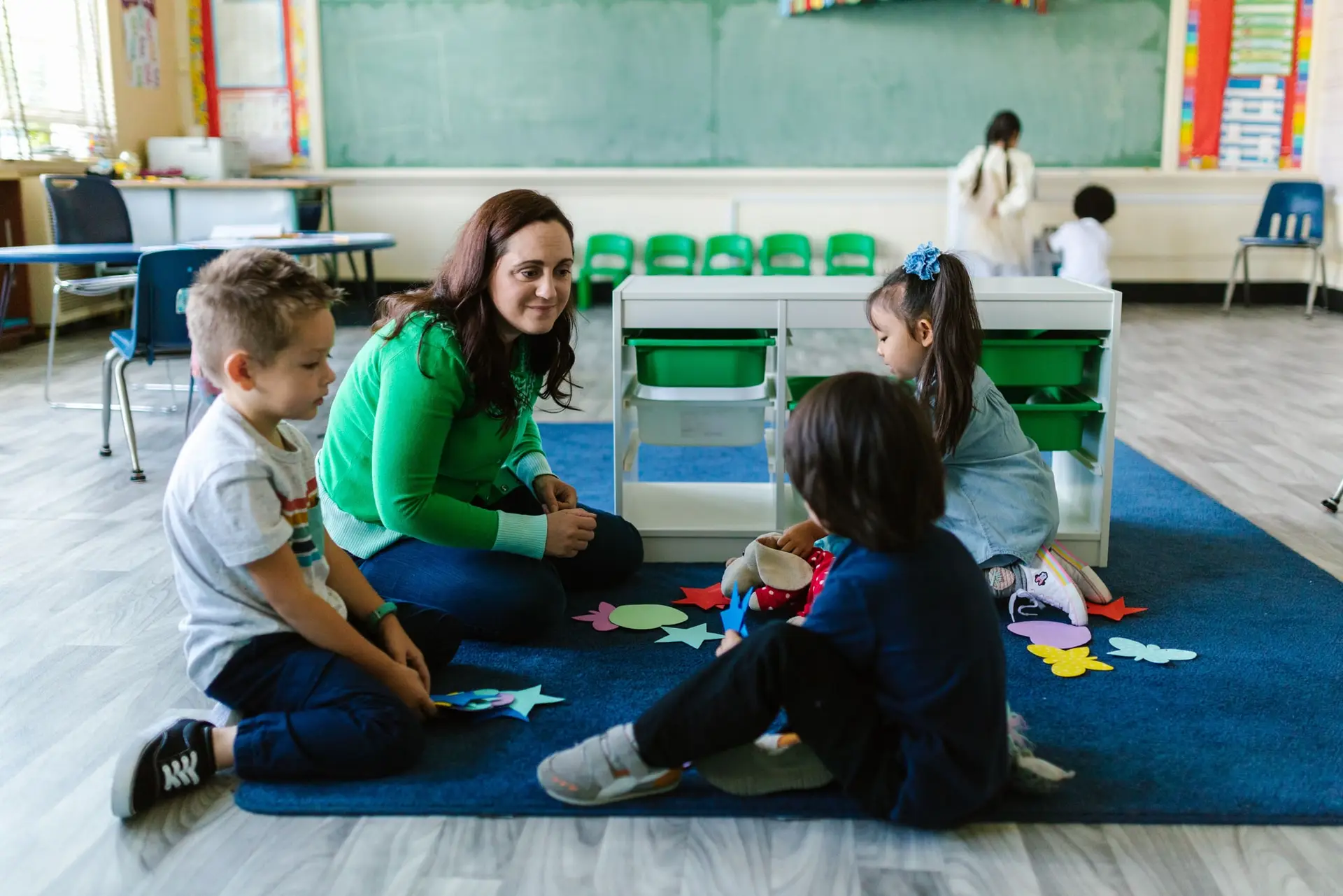 Una docente en el suelo conversa con un grupo de niños sentados sobre una alfombra azul.